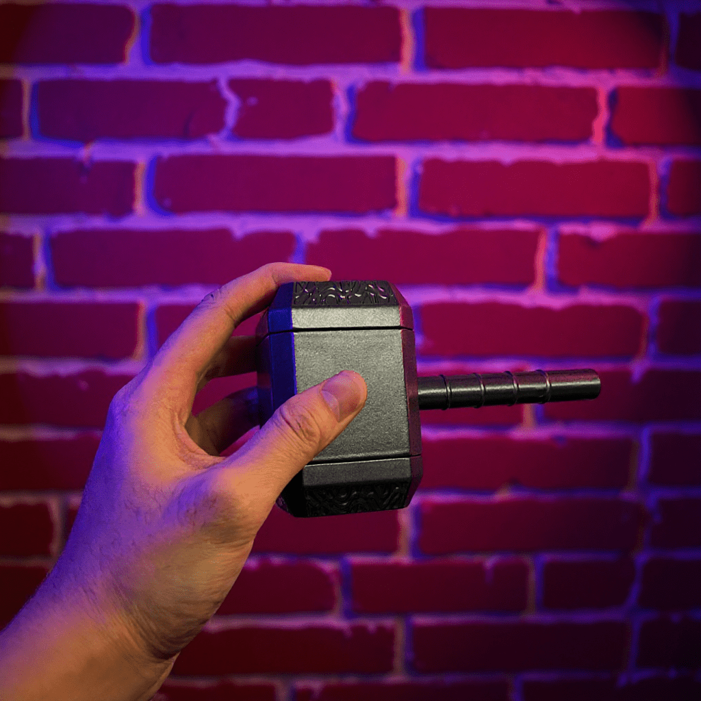 Thor Hammer Shape Grinder – 4 - Part Metal GrinderHand holding a black hammer grinder against a brick wall with purple lighting
