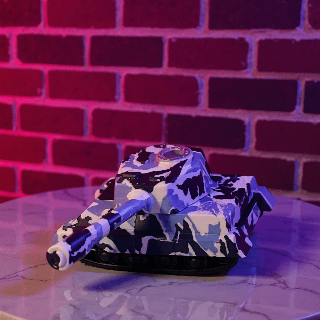 Toy tank grinder with camouflage pattern on a reflective surface in front of a brick wall.