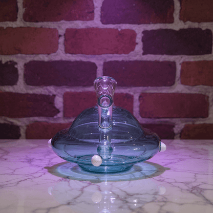 UFO Mini Glass Bong – Water Pipe in 3 Colorsblue glass object on a marble surface with a brick wall background