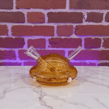 UFO Mini Glass Bong – Water Pipe in 3 ColorsAmber glass pipe with two clear hoses on a marble surface against a brick wall.